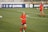 Two female soccer players are on a soccer field wearing red and dark blue uniforms. One player is in the foreground running with a green headband and the number 31 on her shorts, while the other player is in the background. The field has markings and soccer balls are scattered around. Advertising banners with social media icons and text are visible in the background.