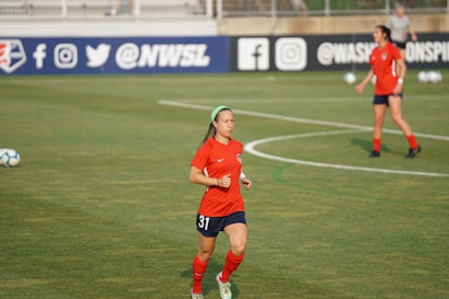Two female soccer players are on a soccer field wearing red and dark blue uniforms. One player is in the foreground running with a green headband and the number 31 on her shorts, while the other player is in the background. The field has markings and soccer balls are scattered around. Advertising banners with social media icons and text are visible in the background.