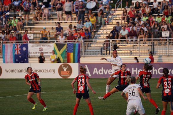A women's soccer match with players actively engaged near the goal area. The player in white is leaping into the air, likely attempting to head the ball, which is visible in the top right. Several players in red and black jerseys are positioned around her, focused on the ball. The background shows a crowd of spectators in the stands, with flags hanging over the railing, including the Australian flag, the Jamaican flag, and a rainbow flag. The atmosphere suggests energetic gameplay.