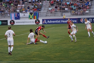 A women's soccer match is taking place on a well-maintained field. One player in a red and black uniform is actively engaged with the ball, while players in white uniforms are attempting to intercept. The game occurs in front of a crowd of spectators seated on bleachers, with banners and flags displayed in the background.