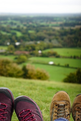 A picturesque view from a hill overlooking a lush green landscape, with two pairs of feet in hiking boots resting in the foreground. The background is slightly blurred, emphasizing the footwear and giving a sense of relaxation.