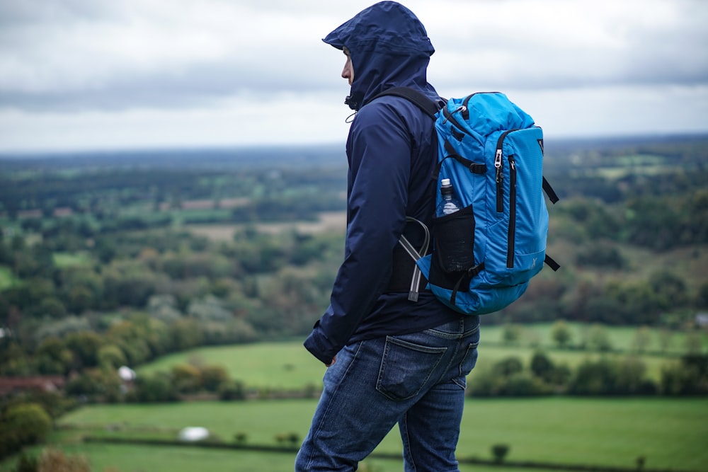 man wearing purple hoodie standing on hill carrying blue backpack