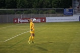 A soccer player dressed in a yellow goalkeeper uniform is performing a kick with the ball balanced on their knee. The setting is a soccer field with advertising banners along the perimeter. In the background, there are trees and a few people sitting in the stands.