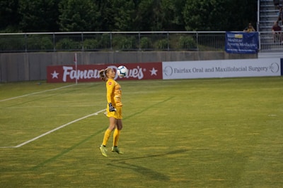 A soccer player dressed in a yellow goalkeeper uniform is performing a kick with the ball balanced on their knee. The setting is a soccer field with advertising banners along the perimeter. In the background, there are trees and a few people sitting in the stands.