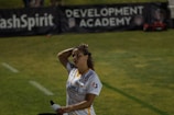 A person in a sports jersey is standing on a field, holding a water bottle and adjusting their hair. The background features signage for a development academy and a section of grass.