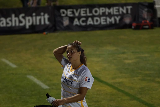 A person in a sports jersey is standing on a field, holding a water bottle and adjusting their hair. The background features signage for a development academy and a section of grass.