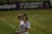 A person in a sports jersey is standing on a field, holding a water bottle and adjusting their hair. The background features signage for a development academy and a section of grass.