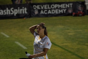 A person in a sports jersey is standing on a field, holding a water bottle and adjusting their hair. The background features signage for a development academy and a section of grass.