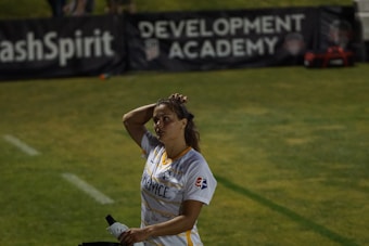 A person in a sports jersey is standing on a field, holding a water bottle and adjusting their hair. The background features signage for a development academy and a section of grass.