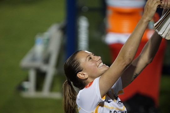 A person with a ponytail, wearing a sports jersey, is smiling broadly and reaching upwards with both hands, possibly holding or grabbing something. The background is blurred, suggesting an outdoor setting, possibly a sports field.