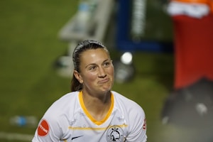 A person wearing a white sports jersey with yellow accents is captured in a candid moment. The expression on their face appears contemplative or perhaps slightly amused. The background is blurred with indistinct shapes and a hint of greenery, suggesting an outdoor setting.