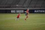 A soccer player and a small child wearing matching red jerseys are on a soccer field. They appear to be running or playing. The field is mostly green, and the background contains stadium seating and advertisements for 'Nike Soccer.'
