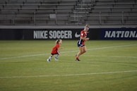 A soccer player and a small child wearing matching red jerseys are on a soccer field. They appear to be running or playing. The field is mostly green, and the background contains stadium seating and advertisements for 'Nike Soccer.'