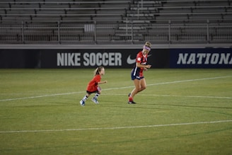 A soccer player and a small child wearing matching red jerseys are on a soccer field. They appear to be running or playing. The field is mostly green, and the background contains stadium seating and advertisements for 'Nike Soccer.'