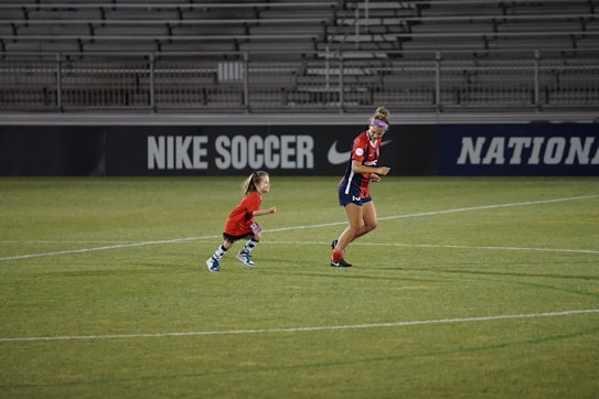 A soccer player and a small child wearing matching red jerseys are on a soccer field. They appear to be running or playing. The field is mostly green, and the background contains stadium seating and advertisements for 'Nike Soccer.'