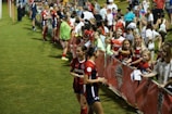 The image features a lively gathering of young soccer players and fans next to a field. Several female athletes in red and blue uniforms interact with an enthusiastic crowd standing behind a barrier. Some people in the crowd are holding objects, like pens and paper, possibly for autographs.