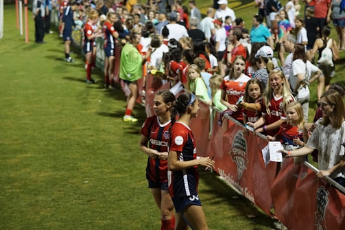 The image features a lively gathering of young soccer players and fans next to a field. Several female athletes in red and blue uniforms interact with an enthusiastic crowd standing behind a barrier. Some people in the crowd are holding objects, like pens and paper, possibly for autographs.