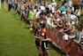 The image features a lively gathering of young soccer players and fans next to a field. Several female athletes in red and blue uniforms interact with an enthusiastic crowd standing behind a barrier. Some people in the crowd are holding objects, like pens and paper, possibly for autographs.