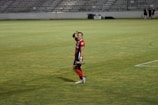 A soccer player in a red and blue uniform waves at the camera while standing on a grassy field. The background features empty bleachers, and the player appears cheerful and relaxed while holding a water bottle.