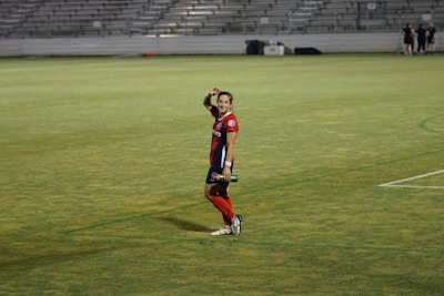 A soccer player in a red and blue uniform waves at the camera while standing on a grassy field. The background features empty bleachers, and the player appears cheerful and relaxed while holding a water bottle.