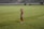 A soccer player in a red and blue uniform waves at the camera while standing on a grassy field. The background features empty bleachers, and the player appears cheerful and relaxed while holding a water bottle.