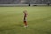 A soccer player in a red and blue uniform waves at the camera while standing on a grassy field. The background features empty bleachers, and the player appears cheerful and relaxed while holding a water bottle.