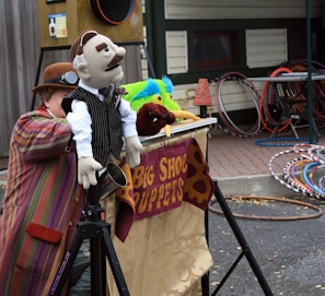 A person dressed in colorful, striped clothing is crouched behind a puppet stage, manipulating hand puppets including one dressed in a suit with a mustache. The stage is decorated with a banner displaying 'Big Shoe Puppets'. Multiple hula hoops are stacked in the background near a wooden building.