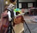 A person dressed in colorful, striped clothing is crouched behind a puppet stage, manipulating hand puppets including one dressed in a suit with a mustache. The stage is decorated with a banner displaying 'Big Shoe Puppets'. Multiple hula hoops are stacked in the background near a wooden building.