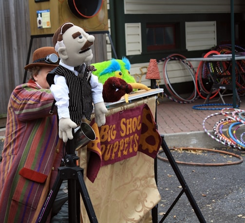 A person dressed in colorful, striped clothing is crouched behind a puppet stage, manipulating hand puppets including one dressed in a suit with a mustache. The stage is decorated with a banner displaying 'Big Shoe Puppets'. Multiple hula hoops are stacked in the background near a wooden building.