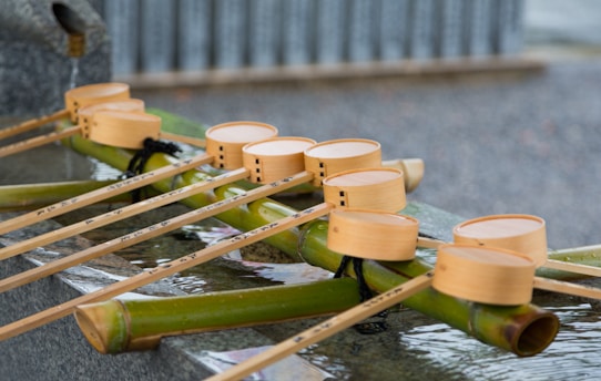 A series of wooden ladles with long handles is placed on a bamboo pipe, with water gently flowing over them. The bamboo is positioned above a stone basin, indicating a traditional Japanese purification fountain, commonly found at the entrance of Shinto shrines.