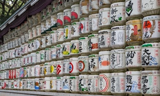 A display of numerous traditional Japanese sake barrels stacked in rows. Each barrel is decorated with colorful and intricate calligraphy and symbols, commonly associated with Japanese culture and rituals. These barrels appear to be part of a ceremonial or decorative installation, often seen at Shinto shrines.