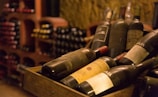 A collection of vintage wine bottles is displayed in a wooden crate within a dimly lit wine cellar. The bottles are dusty, suggesting age and authenticity. In the background, shelves filled with additional bottles create a cozy and rustic atmosphere typical of traditional wine storage areas.