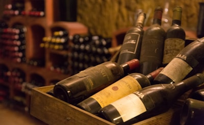 A collection of vintage wine bottles is displayed in a wooden crate within a dimly lit wine cellar. The bottles are dusty, suggesting age and authenticity. In the background, shelves filled with additional bottles create a cozy and rustic atmosphere typical of traditional wine storage areas.