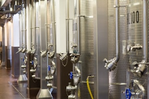 A row of large stainless steel fermentation tanks is lined up in an industrial setting, possibly a brewery or a winery. The tanks have various pipes, valves, and gauges attached to them. The environment appears clean and organized, with industrial flooring and overhead lighting.