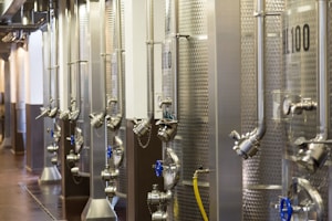 A row of large stainless steel fermentation tanks is lined up in an industrial setting, possibly a brewery or a winery. The tanks have various pipes, valves, and gauges attached to them. The environment appears clean and organized, with industrial flooring and overhead lighting.