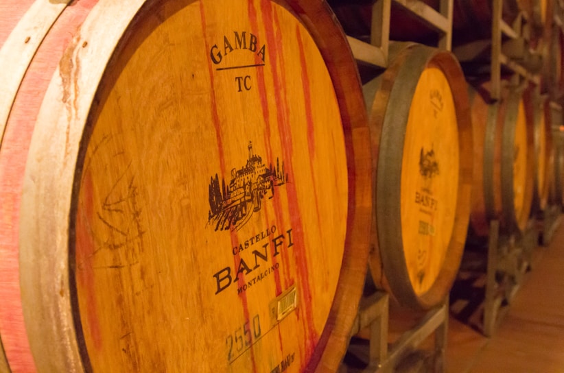 Several wooden wine barrels are stacked horizontally in a dimly lit cellar. Each barrel has inscribed branding, featuring the words 'Castello Banfi Montalcino' and a castle illustration. The barrels have a natural wood tone with red streaks and iron bands securing them.