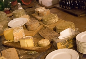 Several rounds of cheese, some with a textured rind and others with a smooth yellow surface, are displayed on wooden boards. Empty white plates are stacked nearby, suggesting a tasting or serving arrangement. The setting appears rustic, with various display shelves and a variety of bottles in the background.