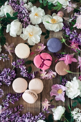 An assortment of pastel-colored macarons is placed on a rustic wooden surface surrounded by a variety of vibrant flowers, including white roses, lilacs, and greenery. The arrangement is artistic and pleasing to the eye.