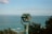 A turquoise binocular viewer stands prominently against the backdrop of a vast ocean landscape. The ocean stretches out to the horizon, with small waves visible and a distant landmass in view. The sky above is clear with sparse clouds, and the scene is bordered by some green foliage.