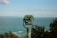 A turquoise binocular viewer stands prominently against the backdrop of a vast ocean landscape. The ocean stretches out to the horizon, with small waves visible and a distant landmass in view. The sky above is clear with sparse clouds, and the scene is bordered by some green foliage.