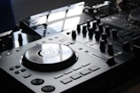 A close-up view of a DJ mixer with a polished surface and multiple dials, buttons, and a large turntable in the center. The equipment is sleek and modern, reflecting light from an overhead source.
