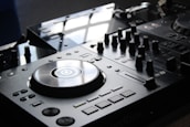 A close-up view of a DJ mixer with a polished surface and multiple dials, buttons, and a large turntable in the center. The equipment is sleek and modern, reflecting light from an overhead source.