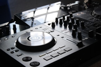 A close-up view of a DJ mixer with a polished surface and multiple dials, buttons, and a large turntable in the center. The equipment is sleek and modern, reflecting light from an overhead source.