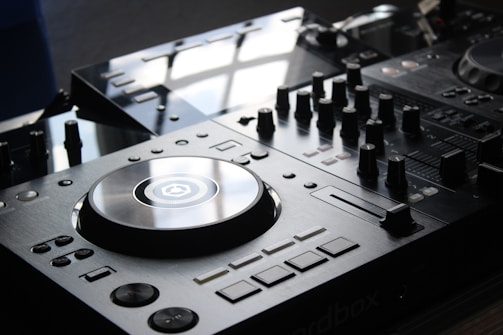 A close-up view of a DJ mixer with a polished surface and multiple dials, buttons, and a large turntable in the center. The equipment is sleek and modern, reflecting light from an overhead source.