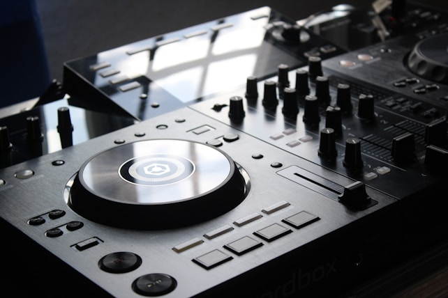 A close-up view of a DJ mixer with a polished surface and multiple dials, buttons, and a large turntable in the center. The equipment is sleek and modern, reflecting light from an overhead source.