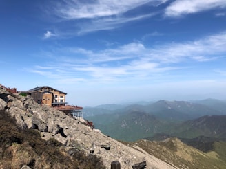 A wooden lodge is situated on a rocky mountainside with a backdrop of distant mountain ranges. The clear blue sky has streaks of white clouds above the scene.