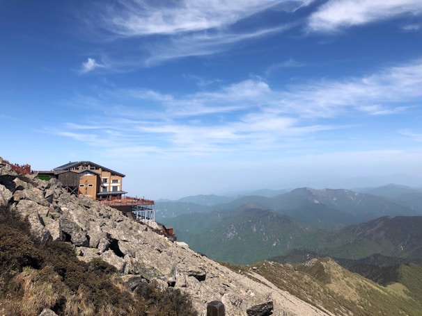 A wooden lodge is situated on a rocky mountainside with a backdrop of distant mountain ranges. The clear blue sky has streaks of white clouds above the scene.