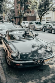 A vintage Alfa Romeo convertible is parked on a street lined with several other vehicles. The car's exterior is shiny with reflections from nearby trees and buildings. In the background, a brick building and some greenery are visible.