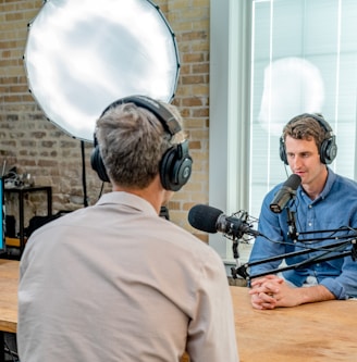 man in gray shirt leaning on table with headphones facing another man leaning on table with headboard