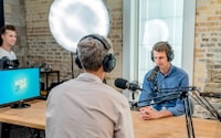 man in gray shirt leaning on table with headphones facing another man leaning on table with headboard